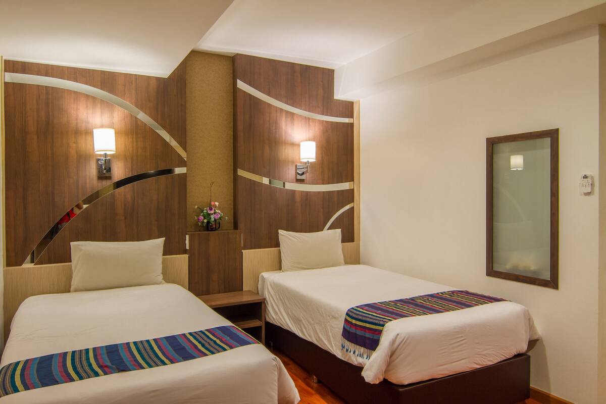 A hotel room with two beds, wooden headboards, wall-mounted lamps, a small table, and a mirror.