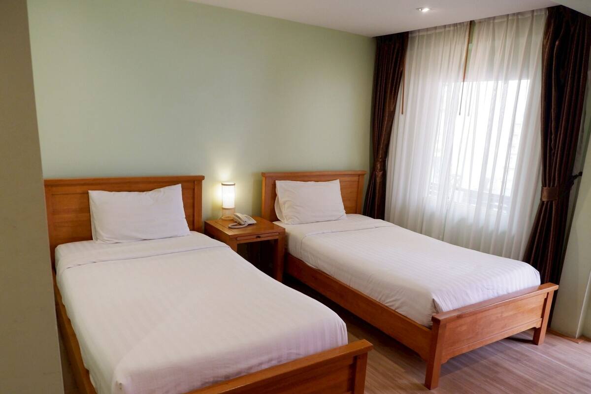 A hotel room with two single beds, wooden headboards, a bedside table with a lamp, and a window with curtains.