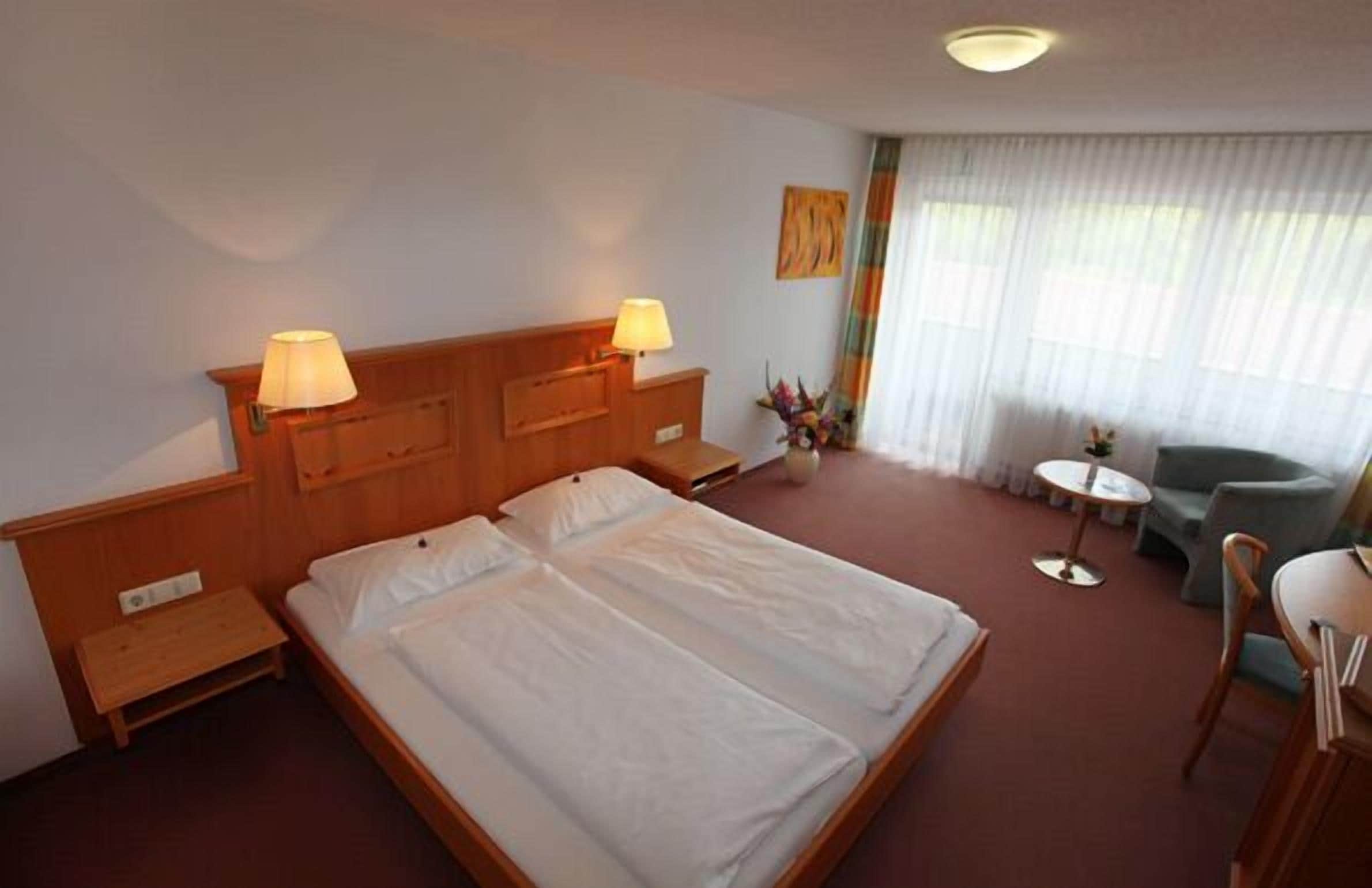 A hotel room with a double bed, two bedside tables with lamps, a small round table with a chair, and a large window with sheer curtains.