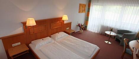 A hotel room with a double bed, two bedside tables with lamps, a small round table with a chair, and a large window with sheer curtains.