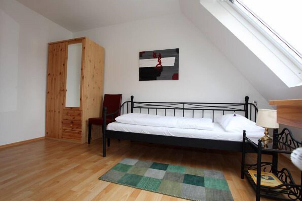 A bedroom with a wooden wardrobe, a bed with white bedding, a green and gray rug, and a painting on the wall.