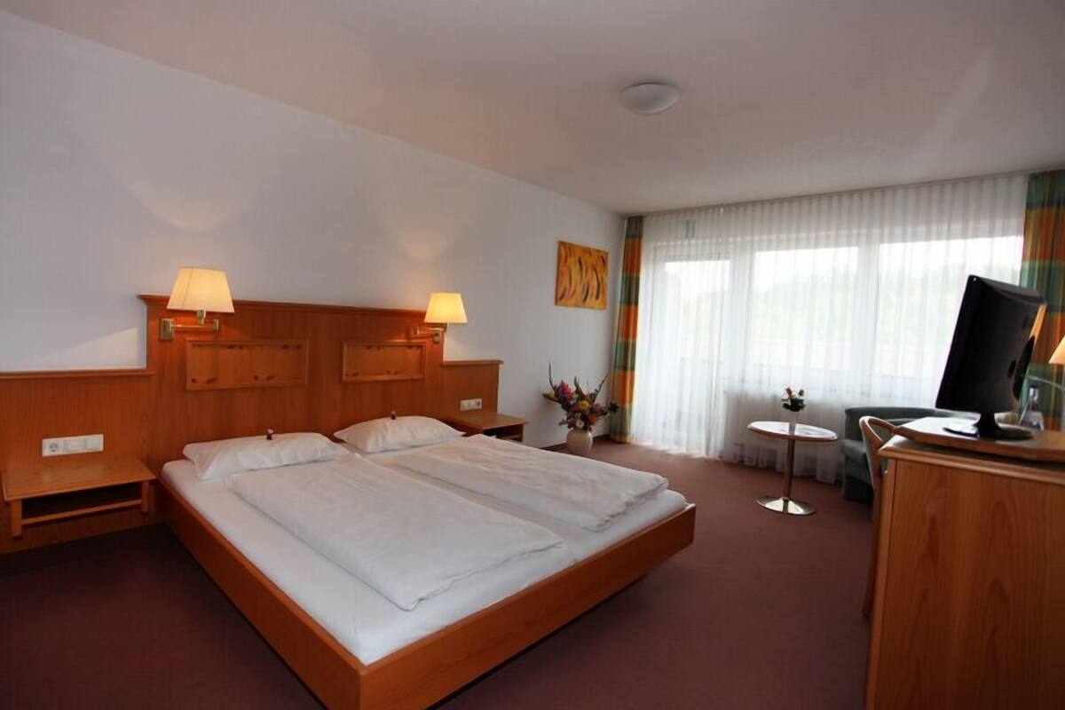 A hotel room with a large bed, two bedside tables with lamps, a small desk with a chair, a TV, and a window with curtains.