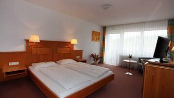 Habitación de hotel con una cama grande, dos mesitas de noche con lámparas, un escritorio pequeño con una silla, un televisor y una ventana con cortinas.