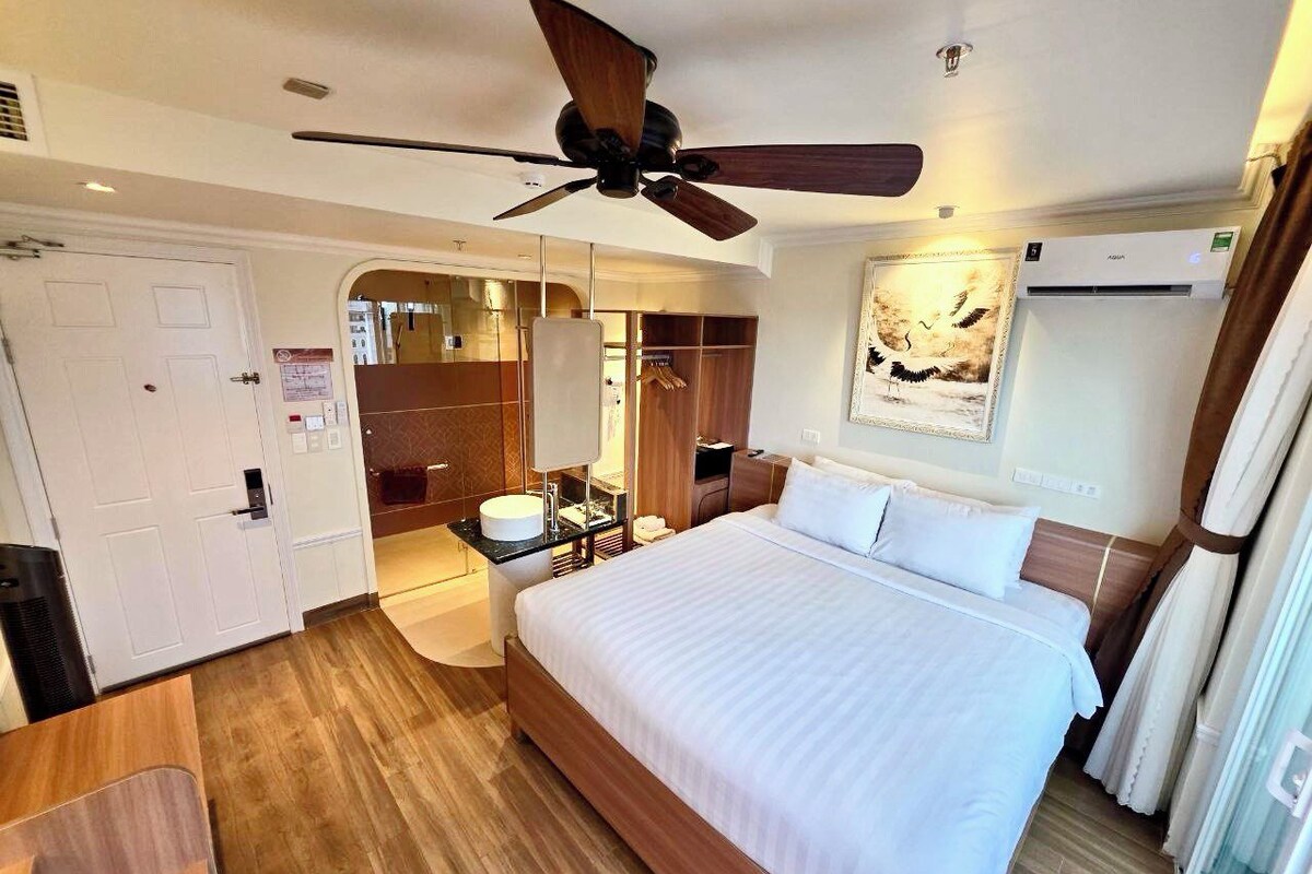 A hotel room with a bed, a ceiling fan, a bathroom with a mirror, and a wardrobe.