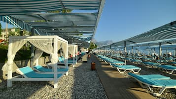 A beachside resort with rows of sun loungers under striped canopies, a wooden walkway, and a clear view of the sea and mountains in the background.