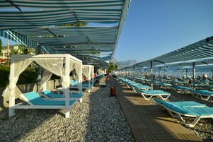 Un resort frente a la playa con hileras de tumbonas bajo toldos a rayas, un paseo de madera y una vista despejada del mar y montañas al fondo.