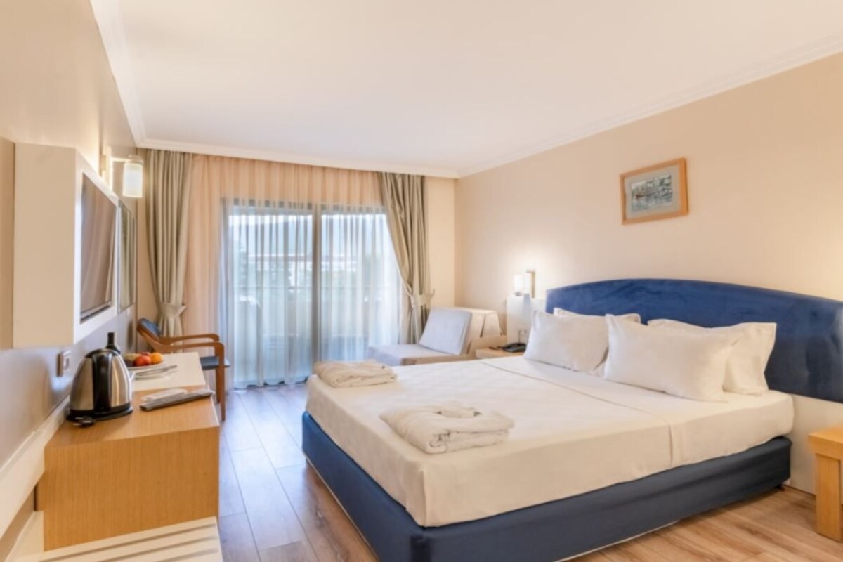 A hotel room with a large bed, a desk with a kettle, a chair, and a bedside table.