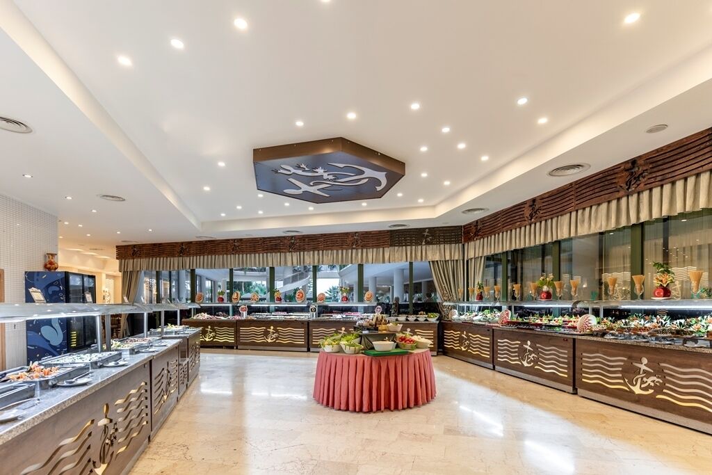 Zona de bufé con varias estaciones de comida, una mesa redonda central con mantel rojo, e iluminación empotrada en el techo con un emblema decorativo.