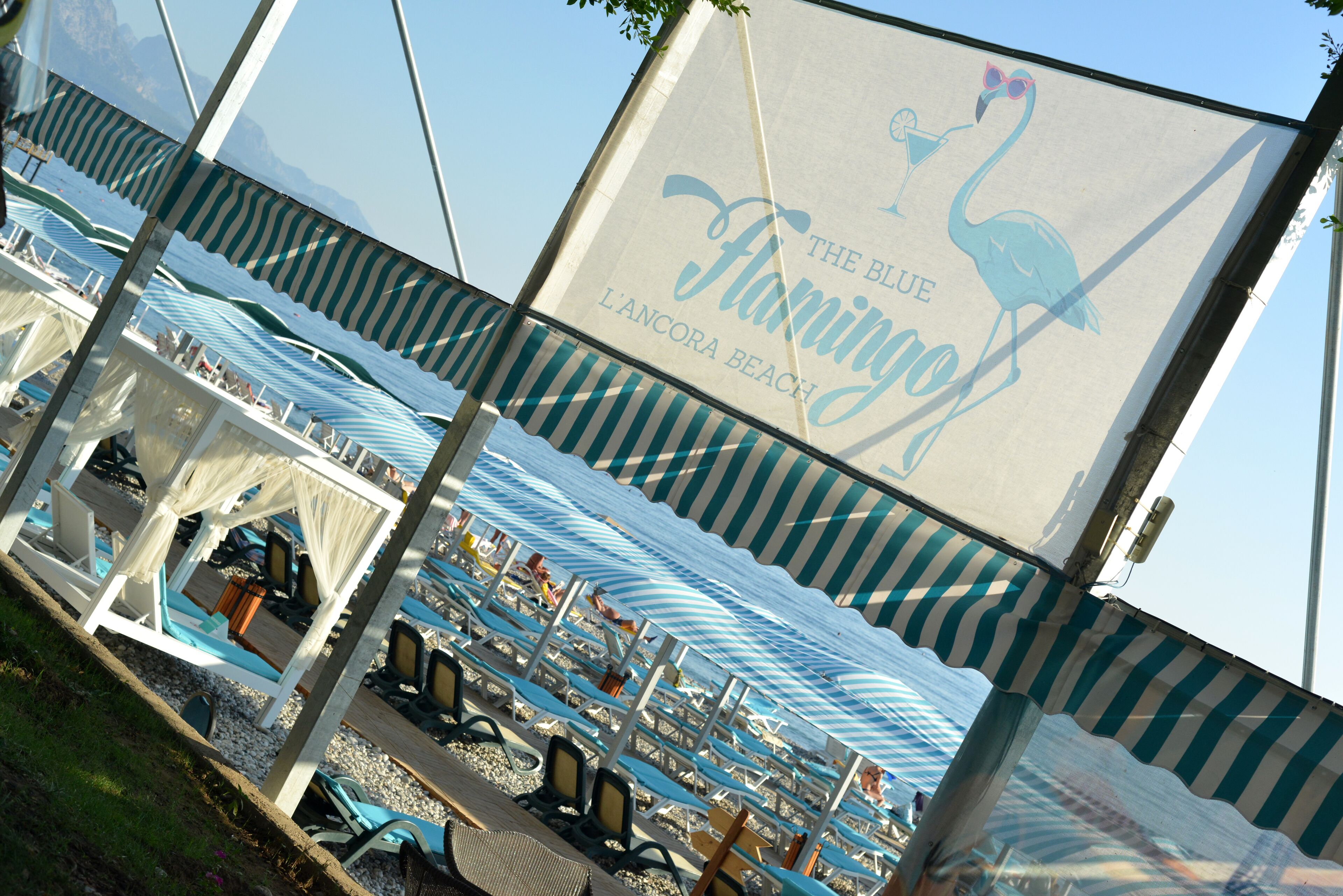 Un resort frente a la playa con sombrillas a rayas y sillas de descanso, con un letrero que dice "The Blue Flamingo L'Ancona Beach".