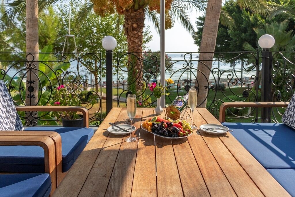 A wooden table with a fruit platter and two glasses of champagne, set on a balcony with blue cushions and a view of the sea.