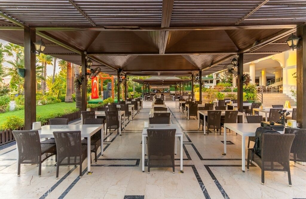 Un espacio exterior para comer con mesas y sillas, cubierto por un techo y rodeado de follaje.