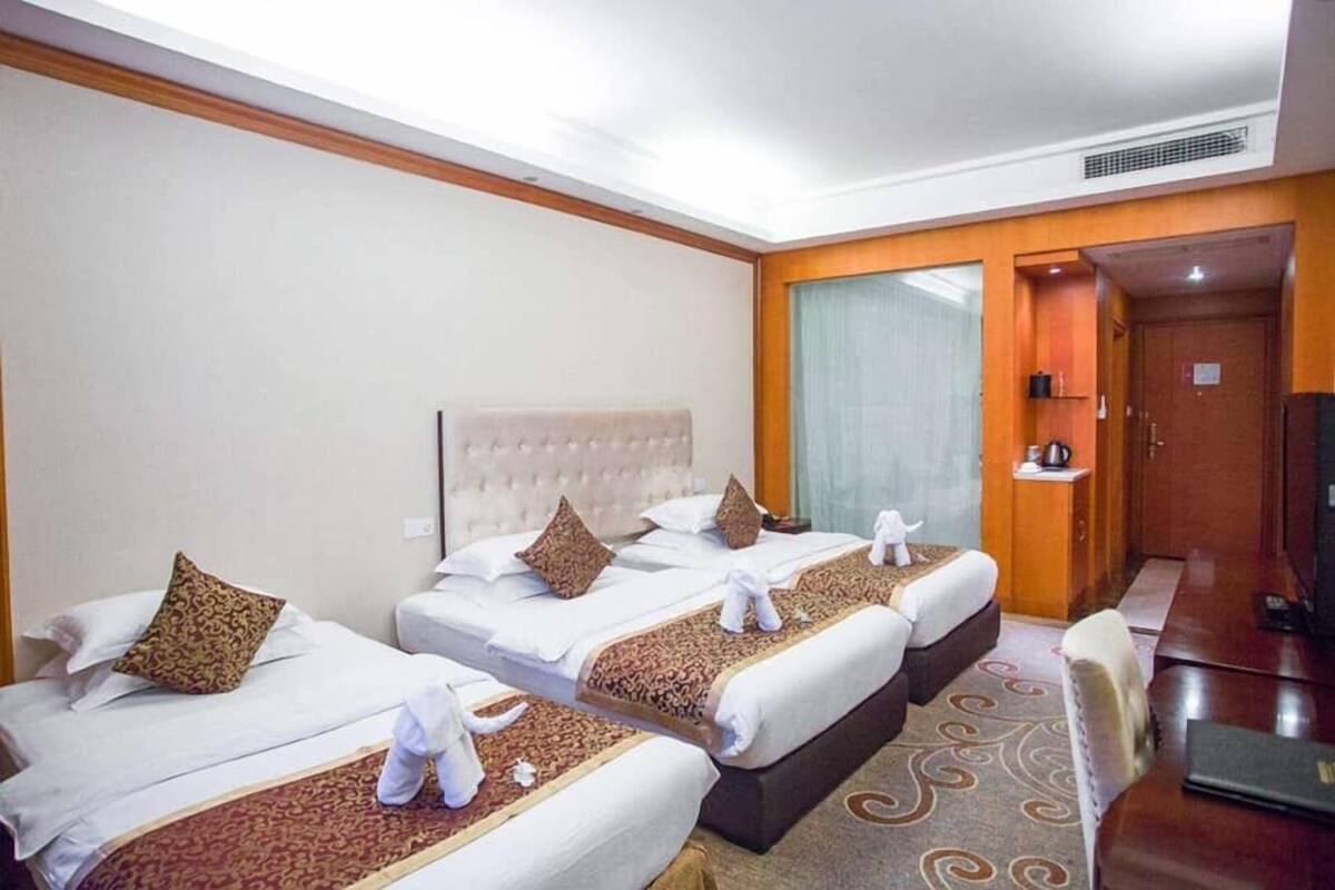 A hotel room with three beds, each with a headboard, white linens, and decorative pillows. There is a desk with a chair and a television.