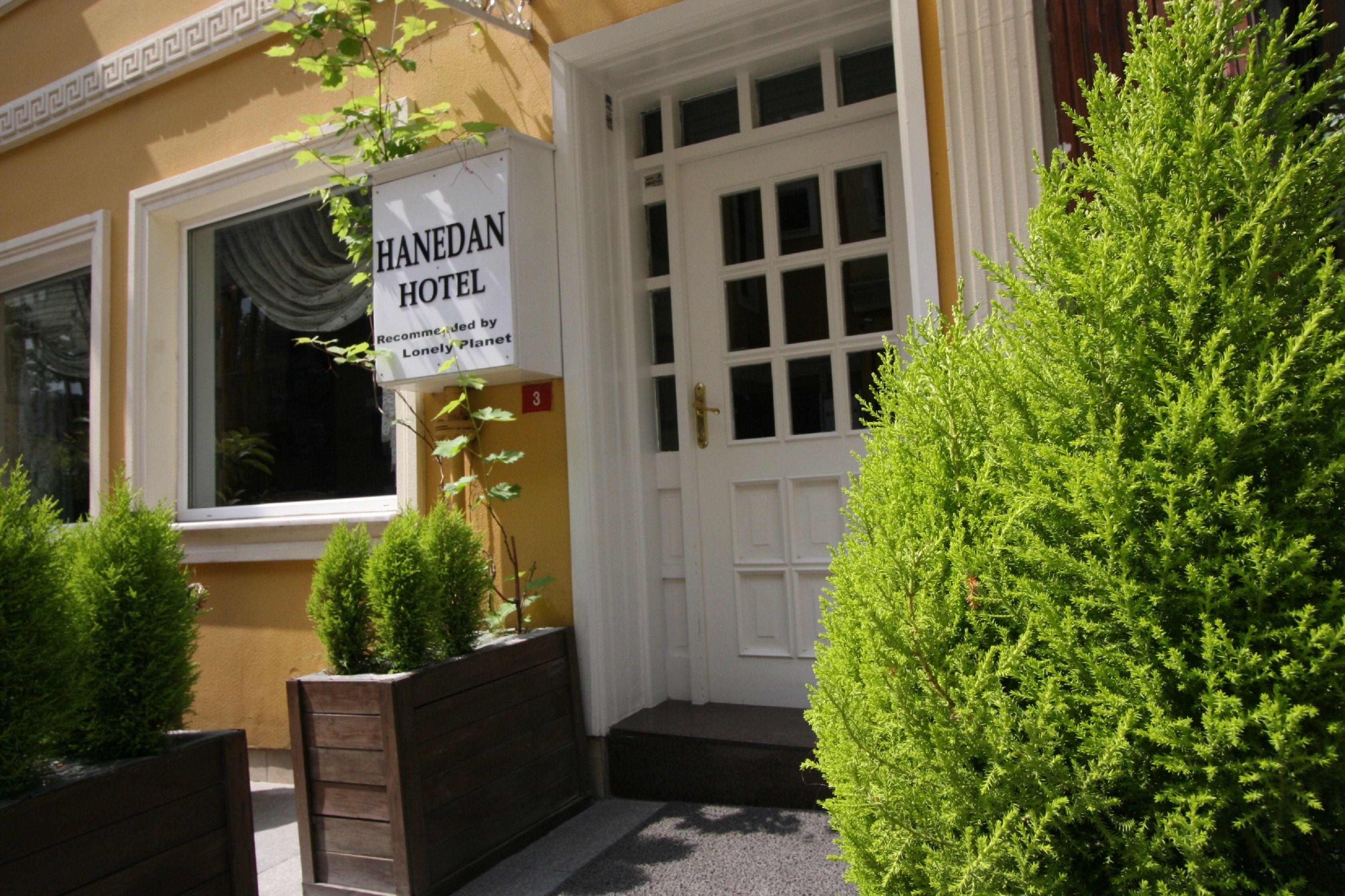 The entrance to Hanedan Hotel, featuring a white door with a grid pattern, a signboard, and neatly trimmed greenery.
