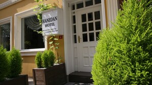 De ingang van Hotel Hanedan, met een witte deur met een rasterpatroon, een bord en verzorgde groene beplanting.