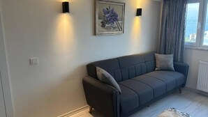 Een moderne woonkamer met een donkergrijze bank, twee wandlampen, een ingelijste schilderij en uitzicht op de zee door een raam met gordijnen.