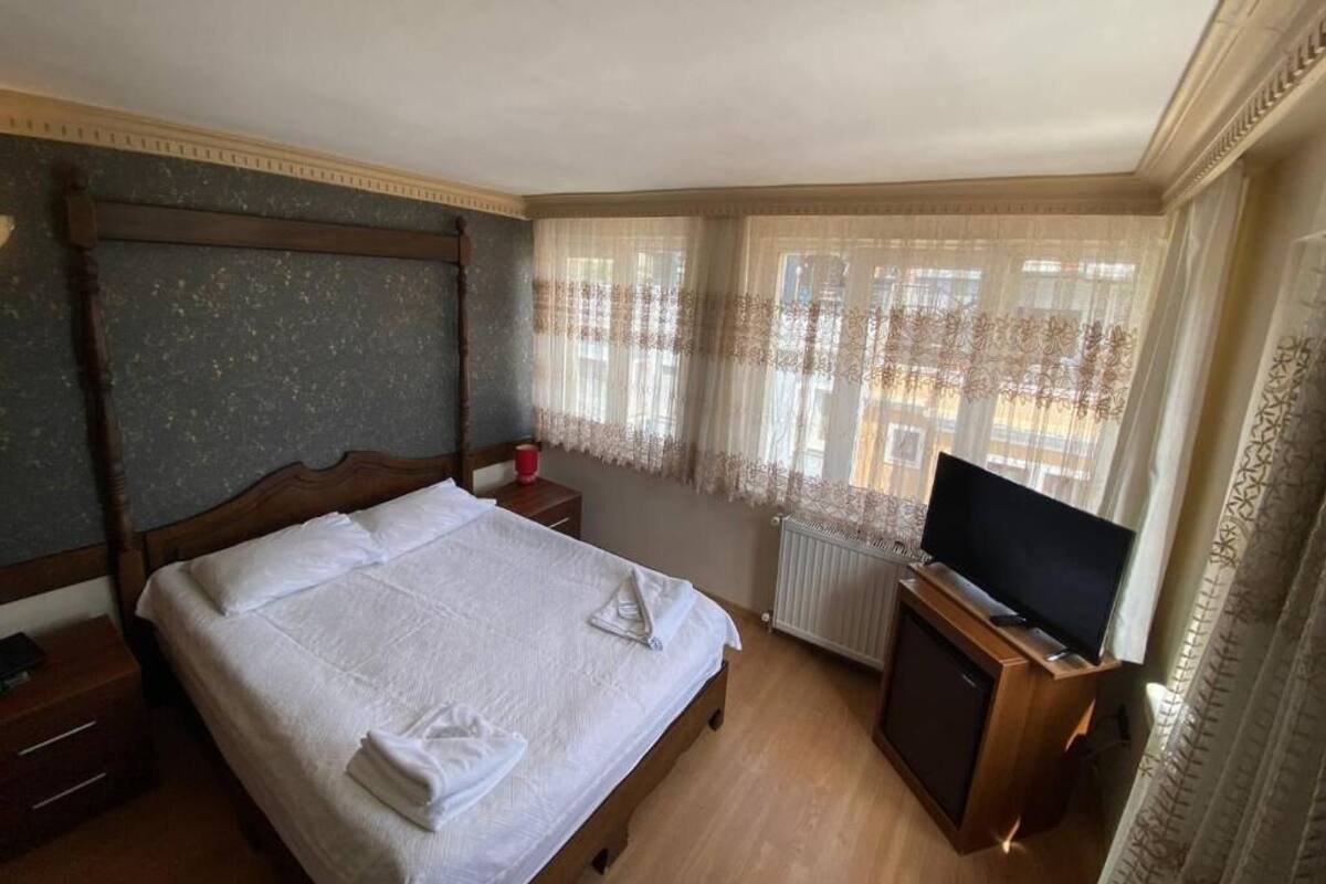 A hotel room with a wooden bed, a television on a wooden stand, a window with sheer curtains, and a radiator.