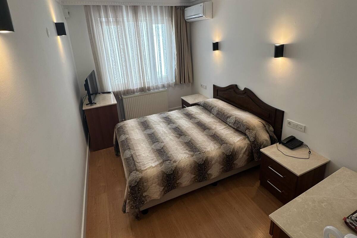 A hotel room with a bed, bedside tables, a desk, a chair, a window with curtains, and an air conditioning unit.