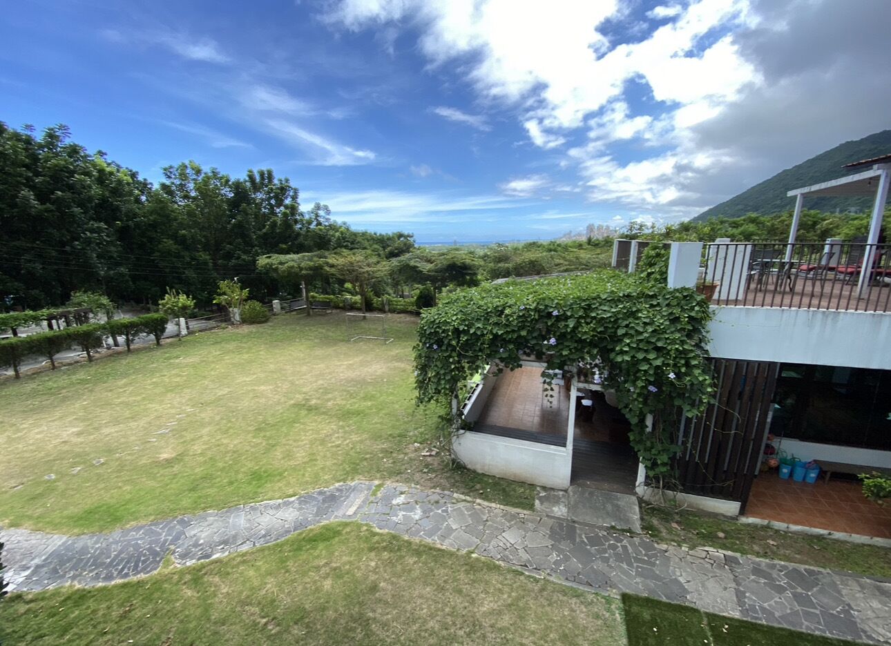 A spacious backyard with a stone pathway leading to a covered seating area, surrounded by lush greenery and a mountainous backdrop.