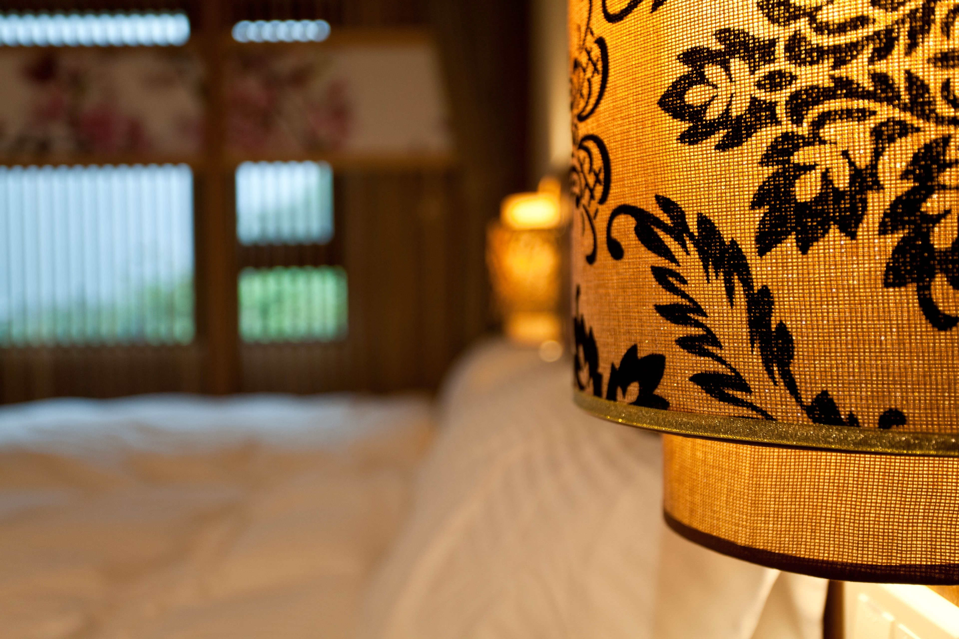 A close-up of a lamp with a floral pattern, a bed with white bedding, and a window with blinds in the background.