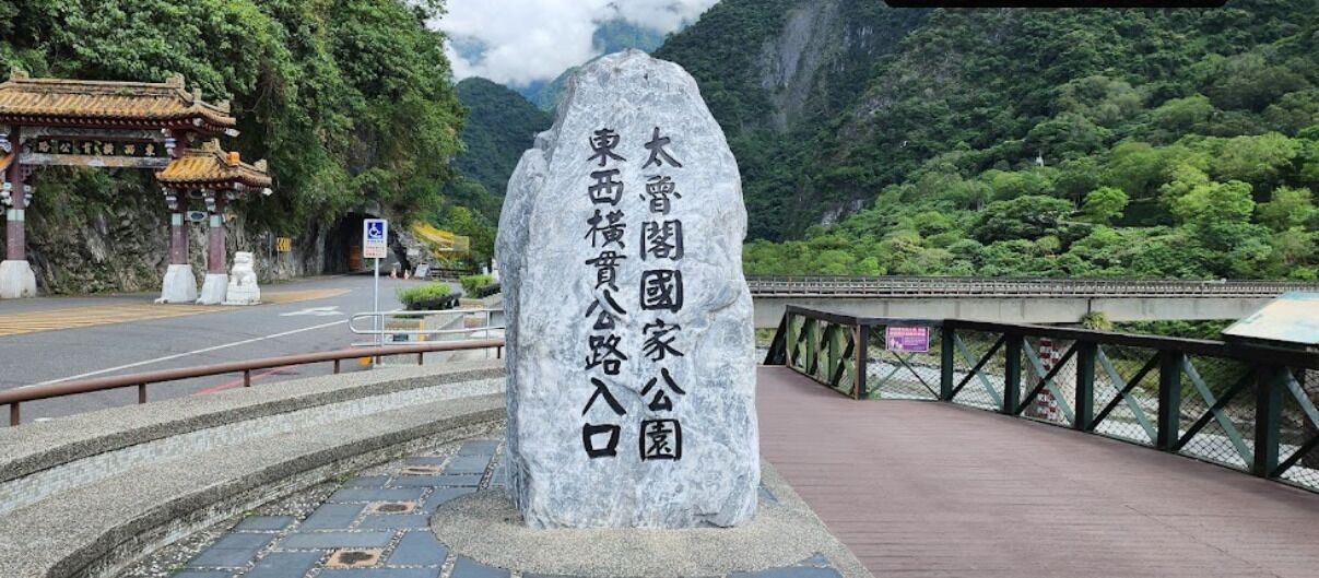 A stone monument with Japanese text, a traditional gate, and a forested mountain backdrop.