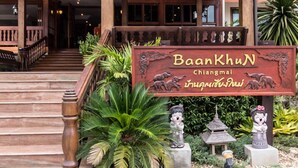 Un escalier en bois mène à un bâtiment sur lequel on peut lire l’inscription « Baan Khun Chiang Mai », et qui est orné de statues d’éléphants.