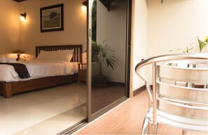 Une chambre avec un lit, une table de chevet, une lampe et une vue sur un balcon accessible par une porte coulissante, où se trouve une chaise.
