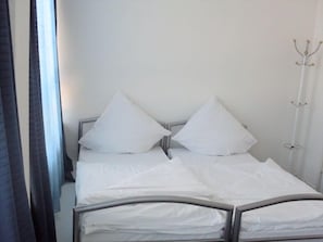 Una cama bien hecha con sábanas y almohadas blancas, cabecera de metal y una lámpara de pie alta y blanca.