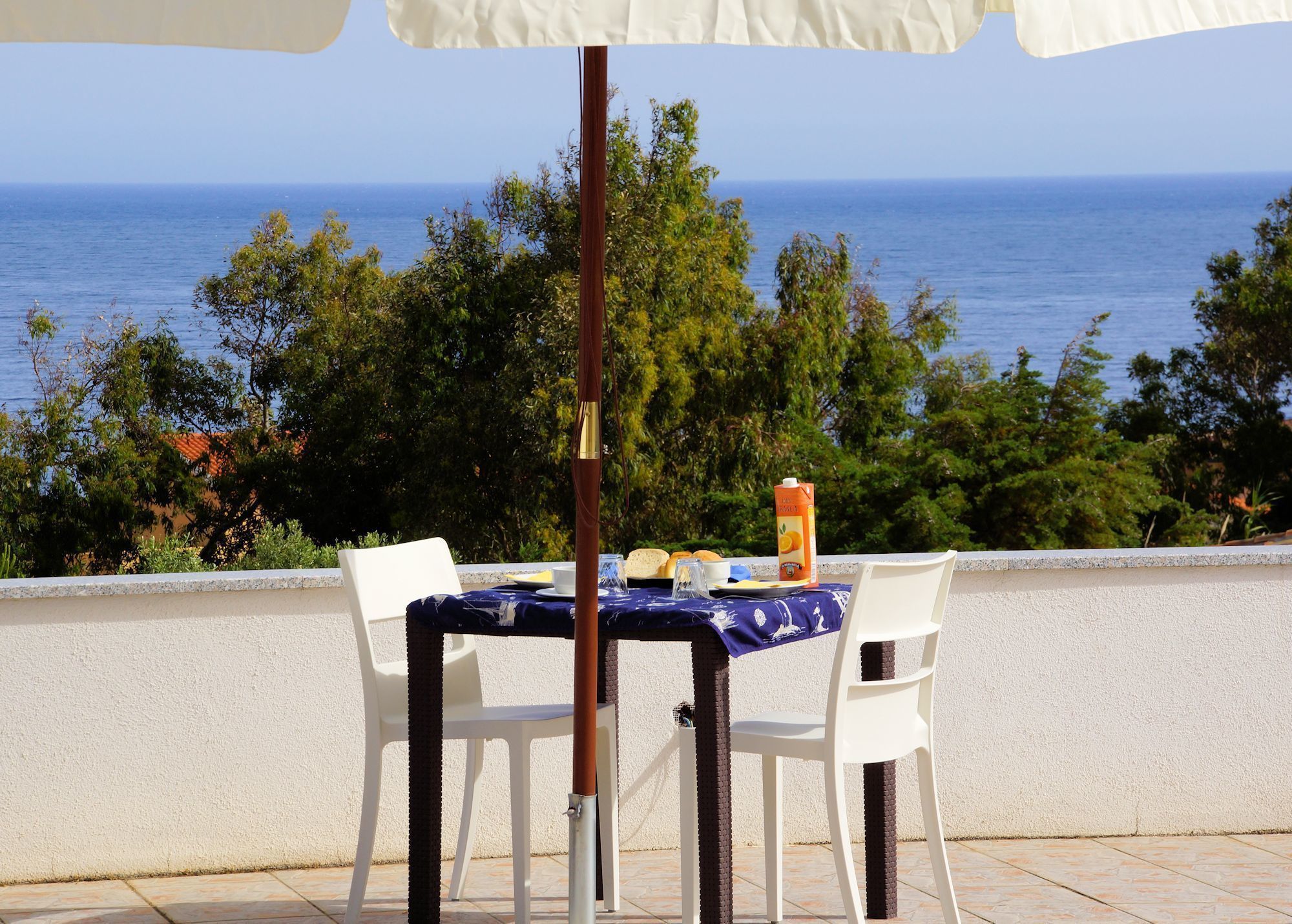 A table with a blue tablecloth and white chairs, a bottle of juice, and a cup, set against a backdrop of a clear blue sky and the sea.