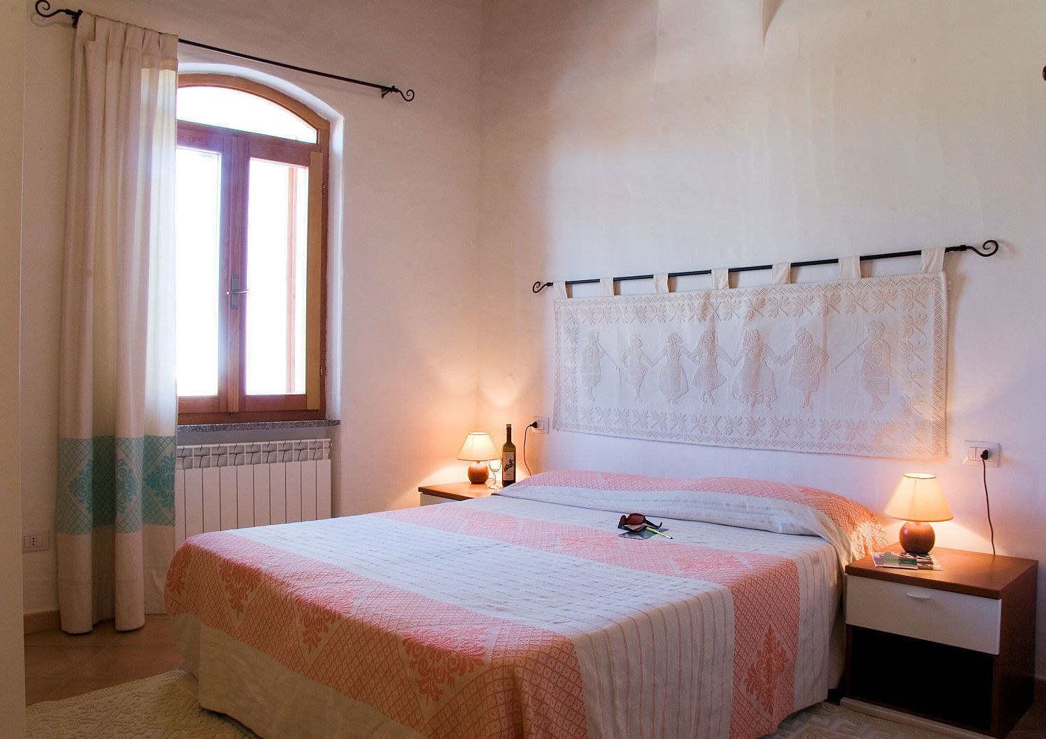 A bedroom with a bed, two bedside tables, a window with curtains, a radiator, and a wall tapestry.