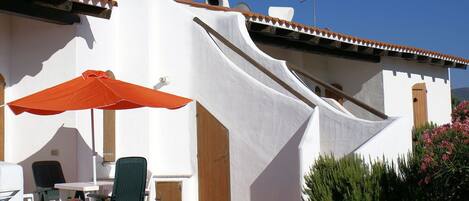 Uma área de estar externa com mesa e cadeiras, um guarda-sol vermelho e um edifício branco com telhado inclinado.