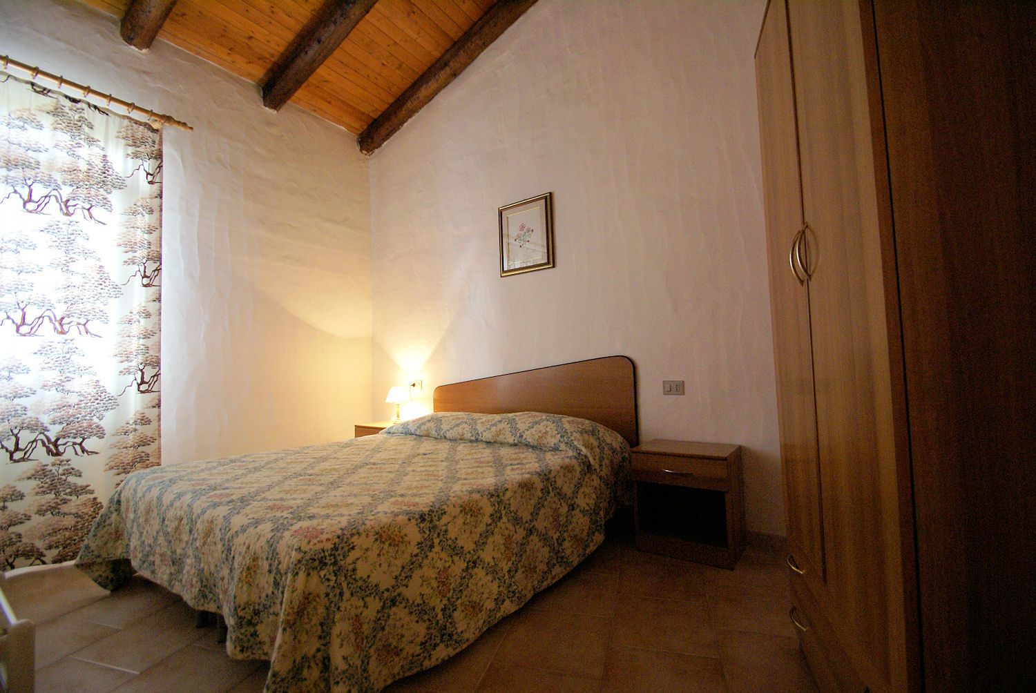 A bedroom with a bed, a nightstand, a wooden wardrobe, and a wall-mounted picture.