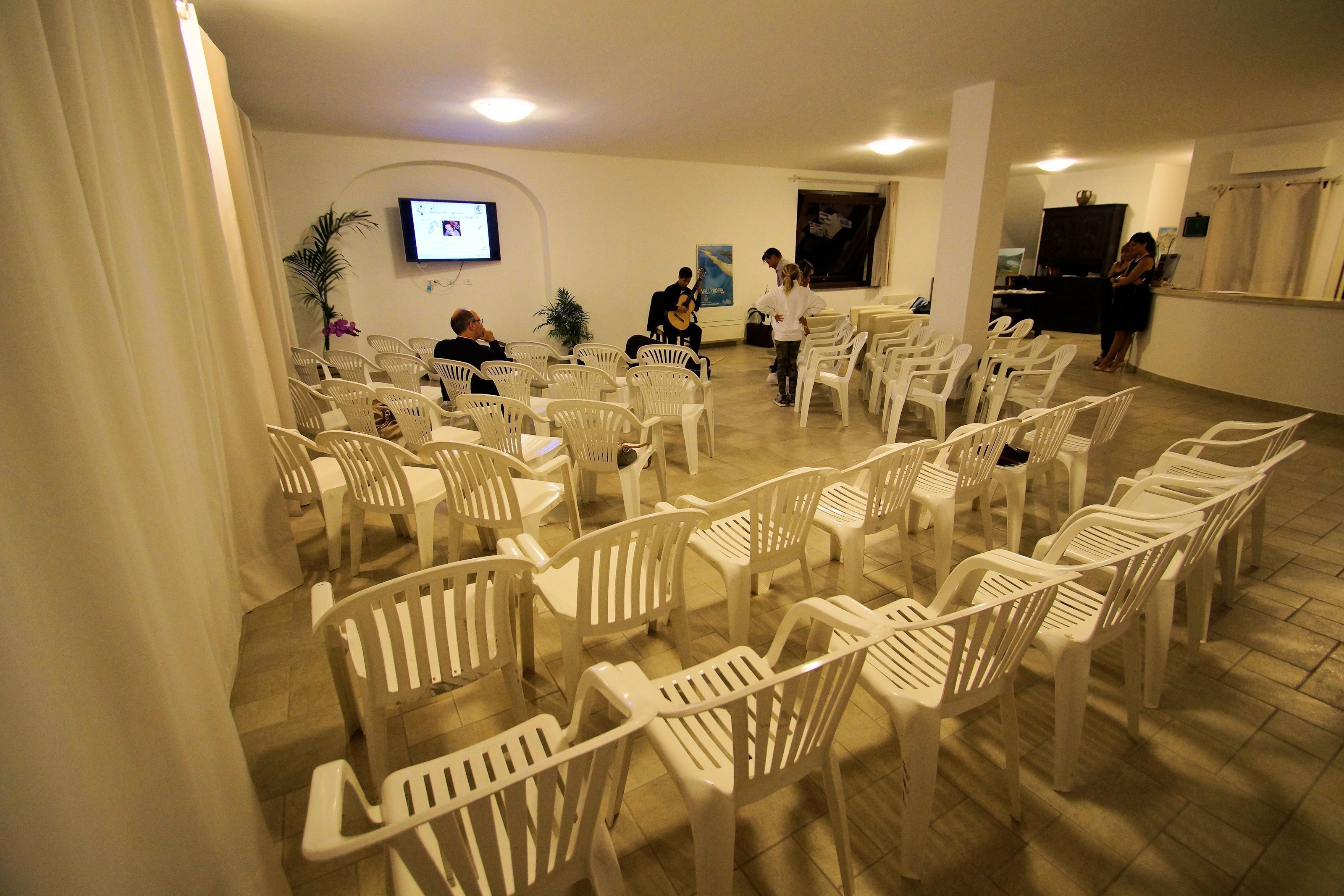 A room with rows of white plastic chairs arranged for an event. A television is mounted on the wall, and a few people are standing near the front.