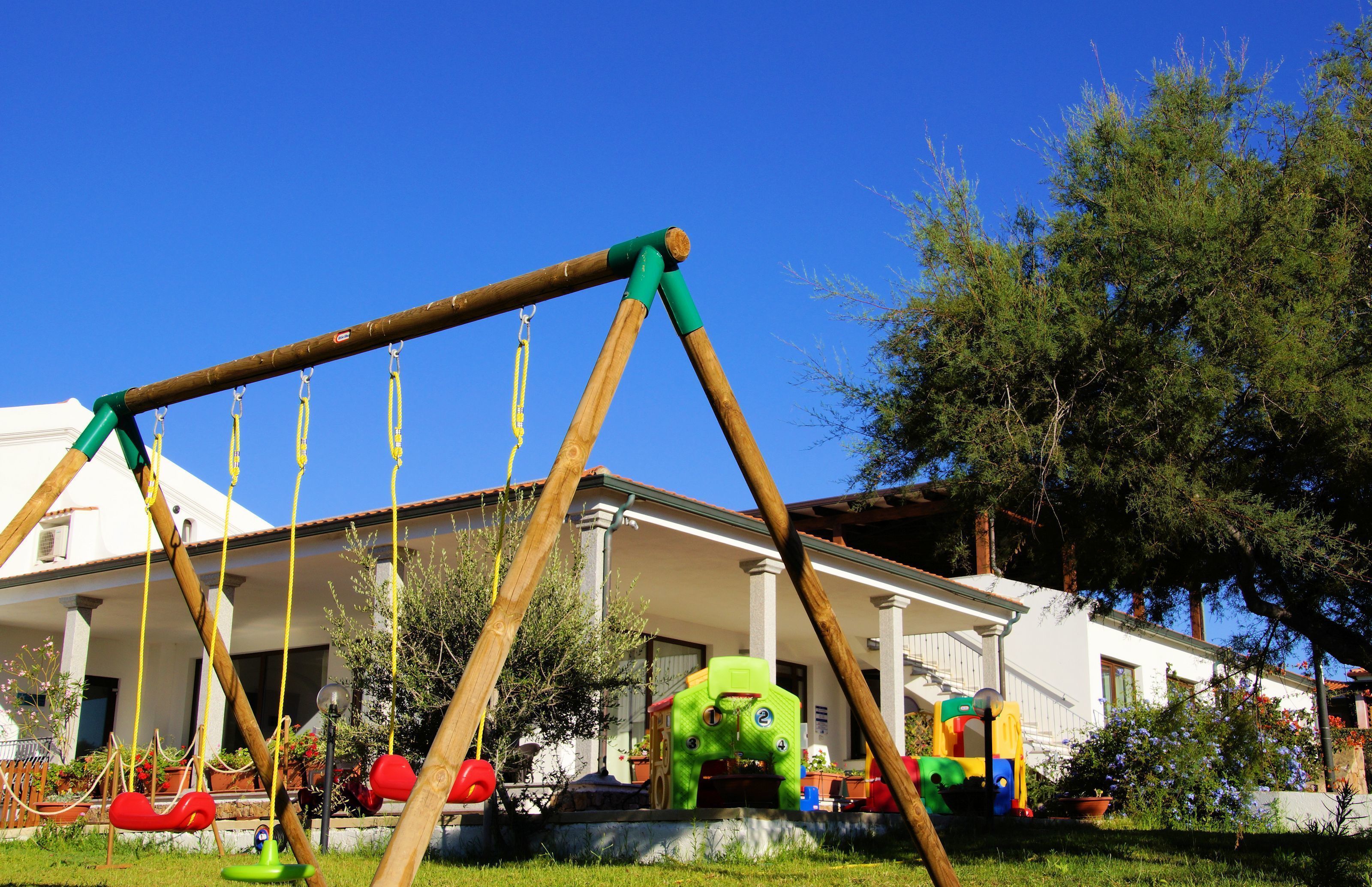 A wooden swing set with multiple swings, a green play structure, and a white building with columns in the background.