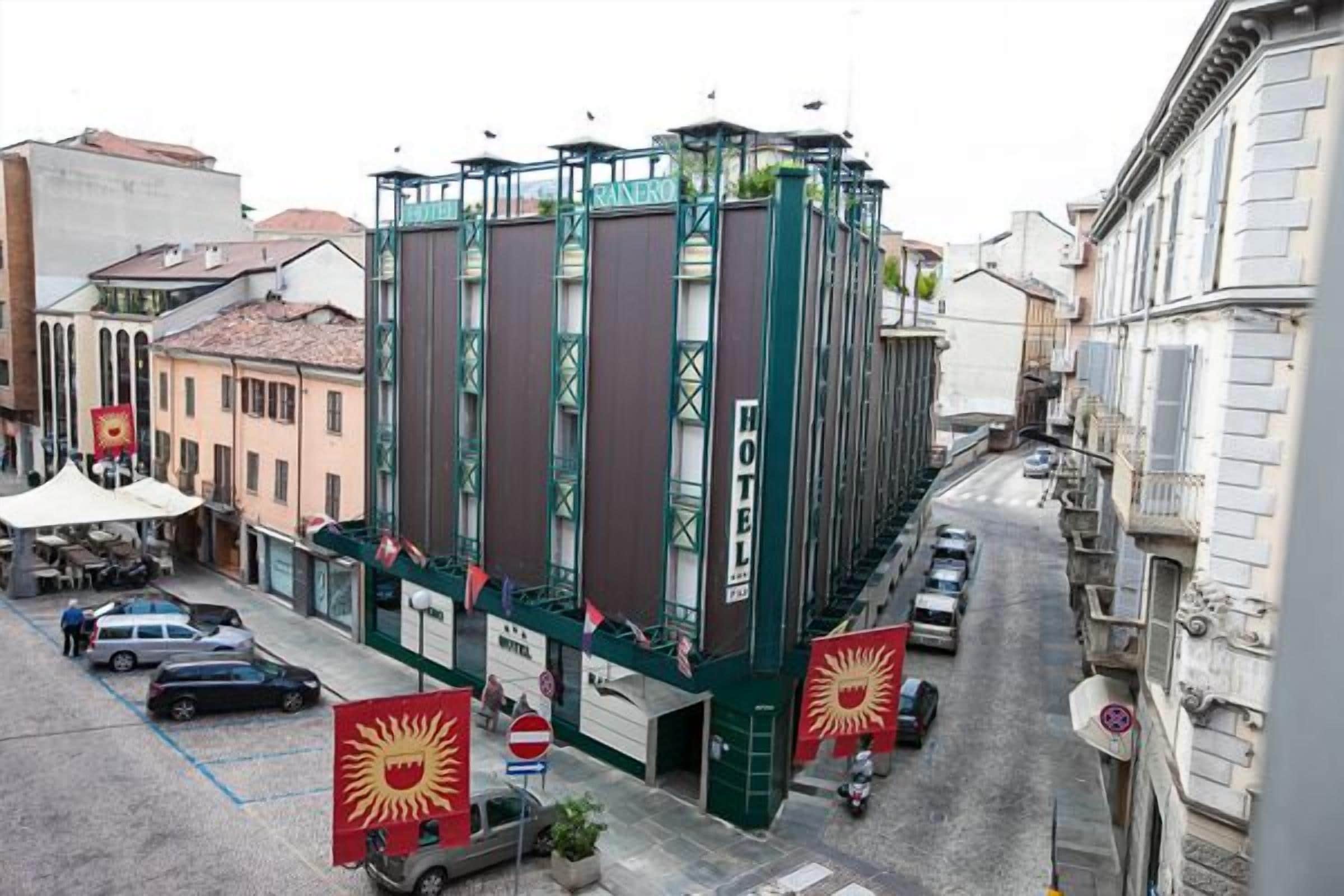 Un hotel con fachada verde, varios pisos y un cartel que indica que es un hotel. Se ven coches aparcados y una acera.