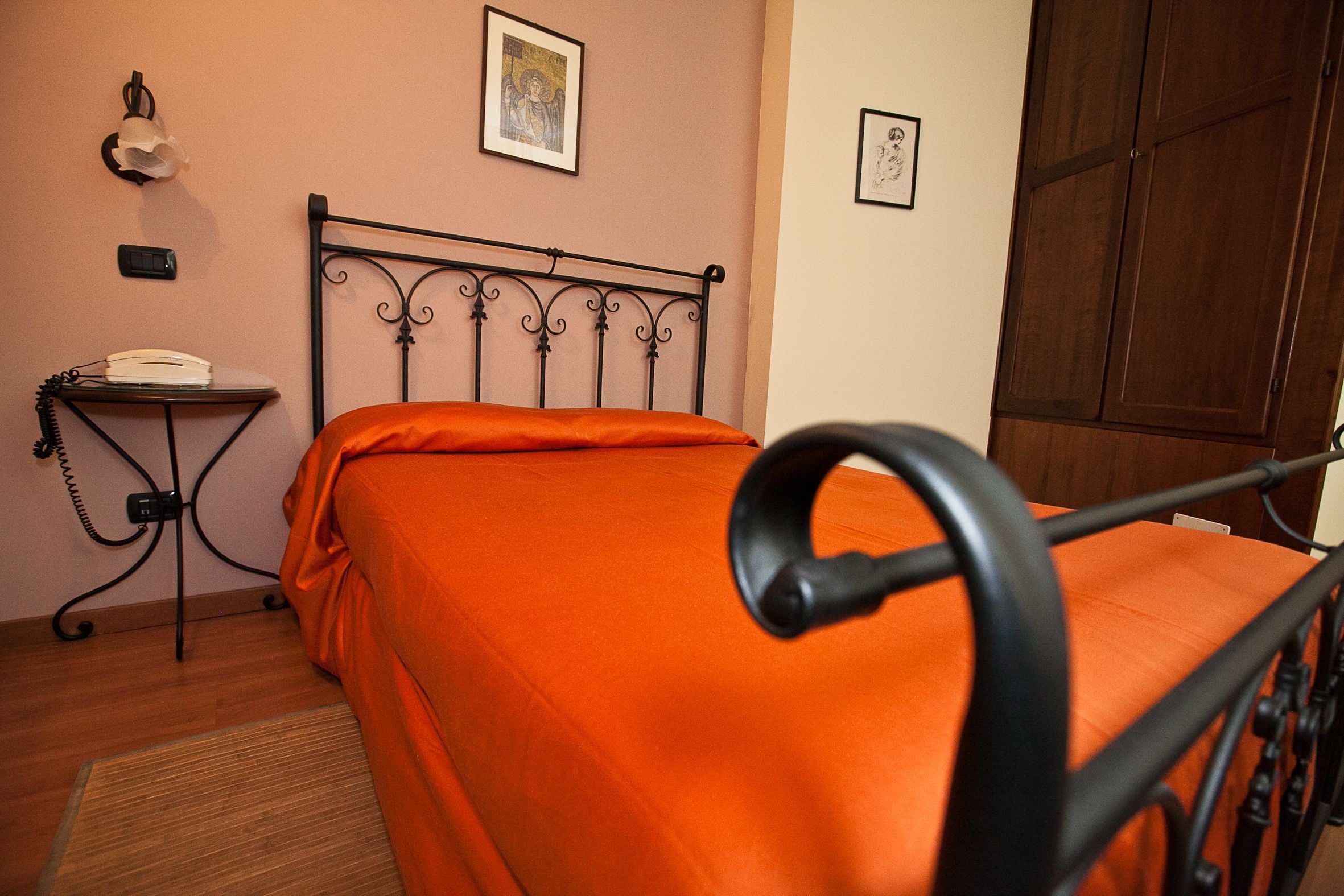 A bedroom with a metal bed frame, an orange bedspread, a bedside table with a lamp, and a wooden wardrobe.