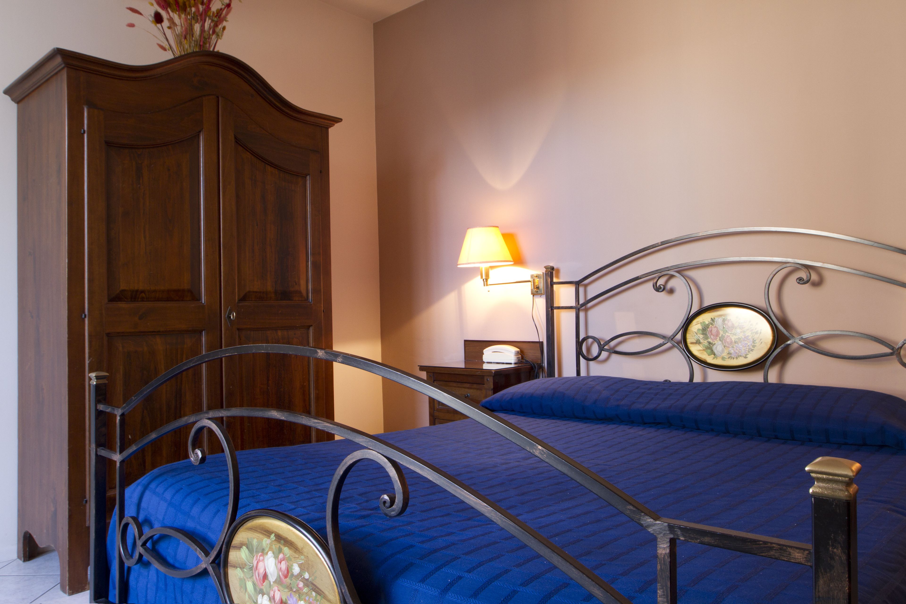 A bedroom with a bed, a wooden wardrobe, a nightstand with a lamp, and a decorative mirror.