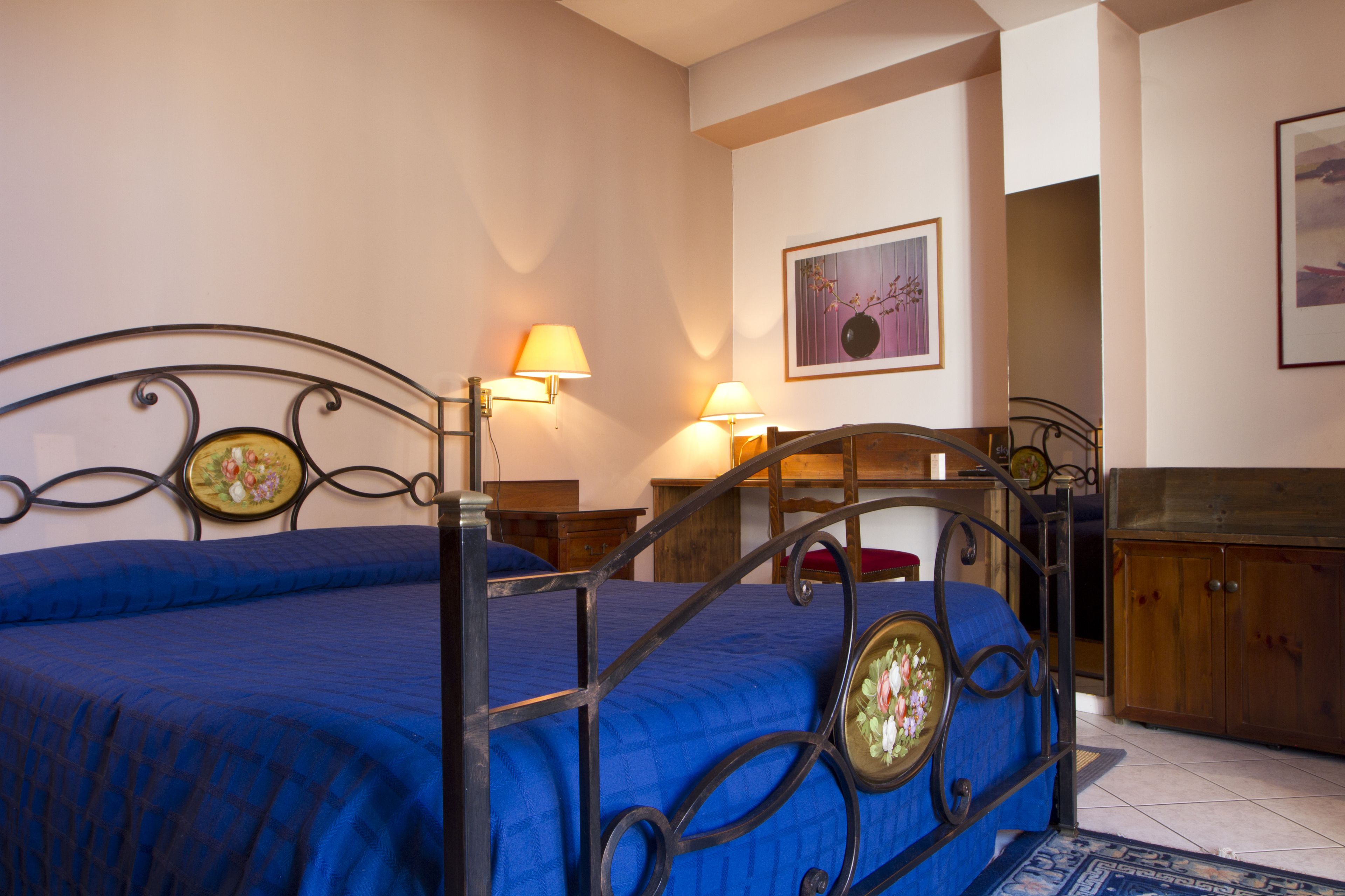 A bedroom with a metal bed frame, a blue bedspread, two bedside lamps, a wooden nightstand, and a wooden cabinet.