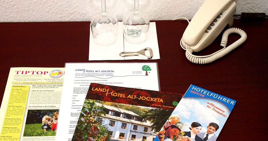 A desk with a telephone, a brochure titled "LAND HOTEL ALT-JOCKETA," a menu, and a glass with a bottle.