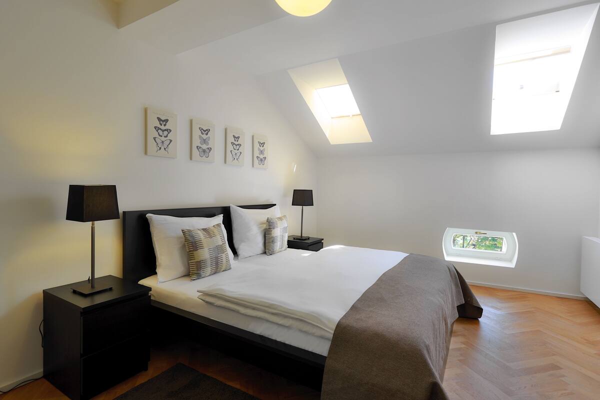 A bedroom with a bed, two bedside tables, a window, and skylights.