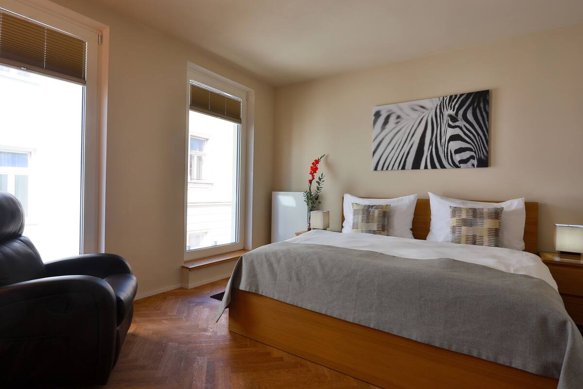A hotel room with a large bed, a massage chair, a window with blinds, and a zebra artwork on the wall.