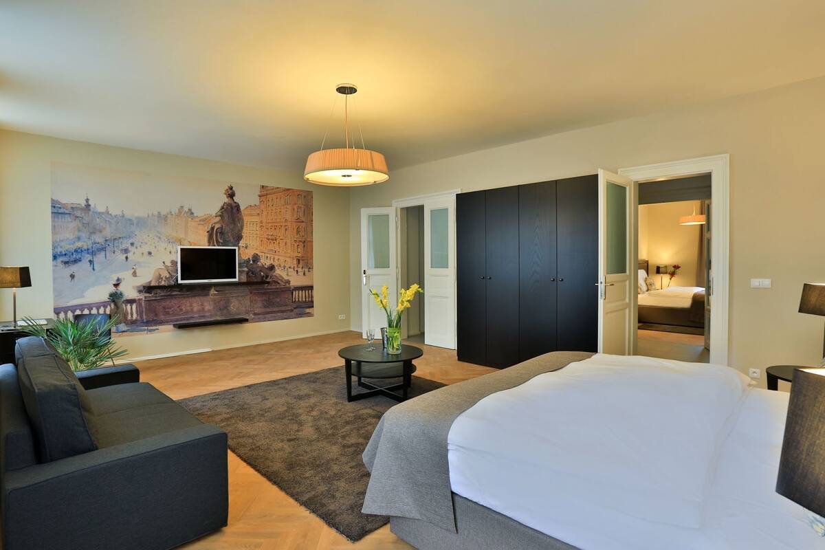 A modern hotel room with a bed, a sofa, a TV, a painting, and a door leading to another room.