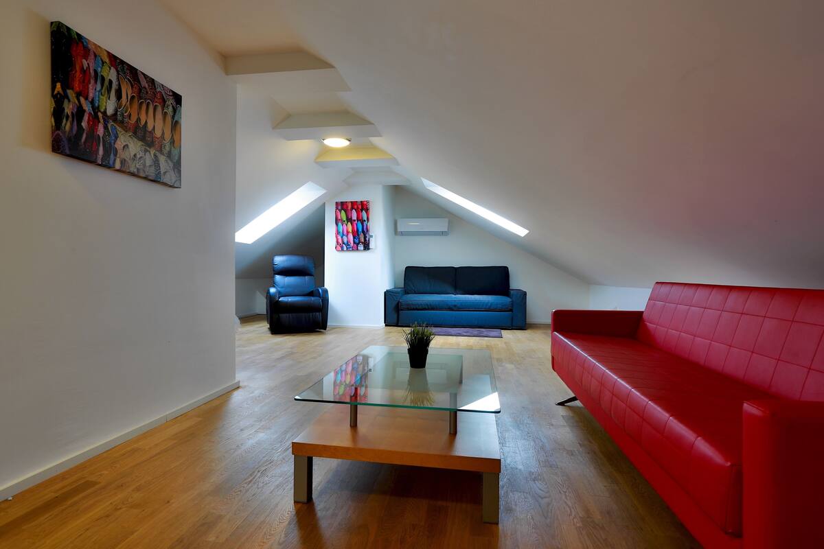 An attic living room with a glass coffee table, red sofa, blue sofa, and a colorful painting on the wall.