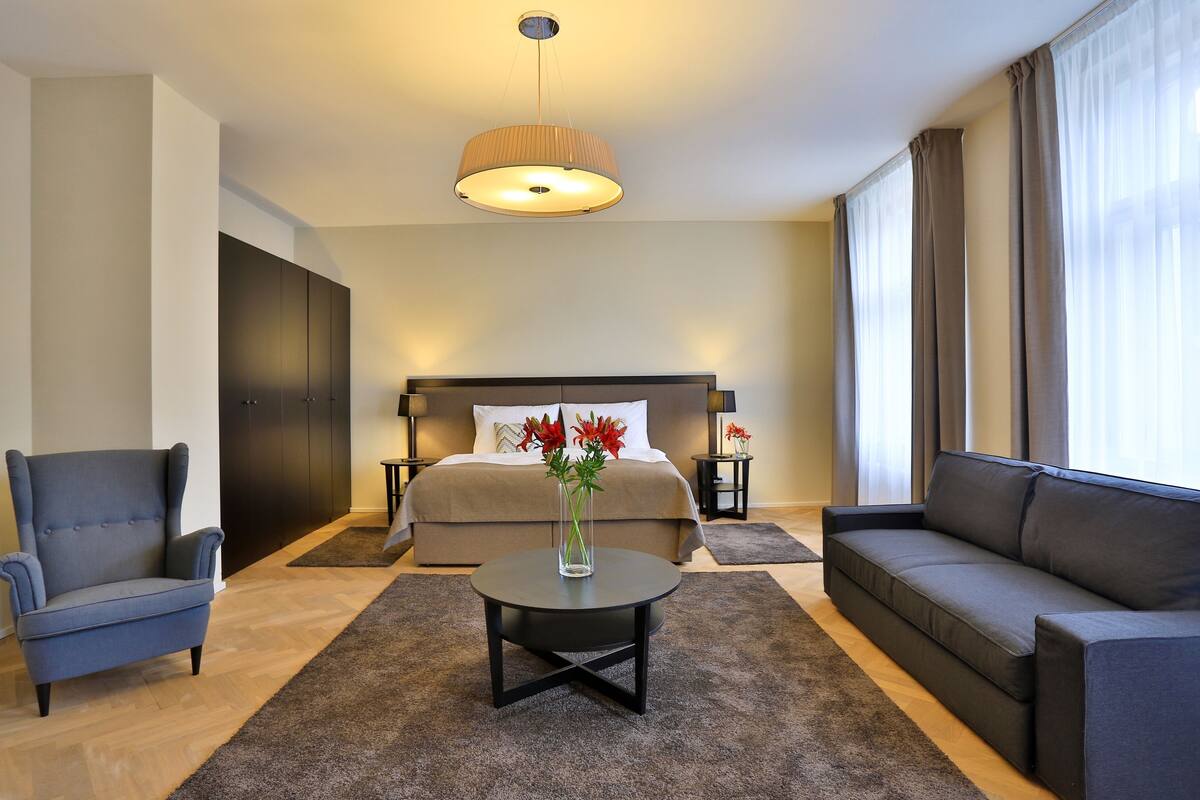 A modern hotel room with a bed, a sofa, a chair, a coffee table, and a vase with flowers.
