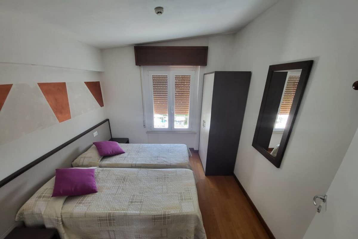 A bedroom with two beds, a window with blinds, a wardrobe, and a mirror on the wall.