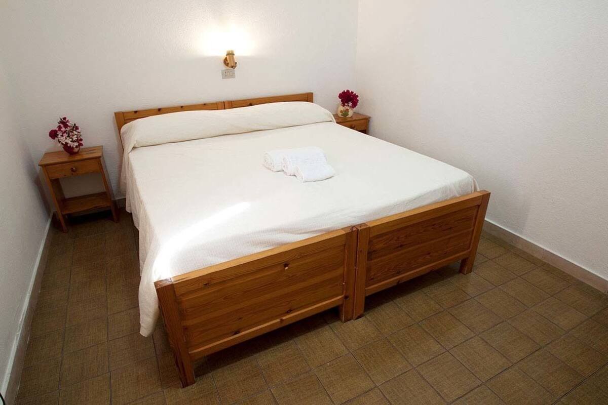 A single bed with a wooden frame, white linens, and a small bedside table with a vase of flowers.