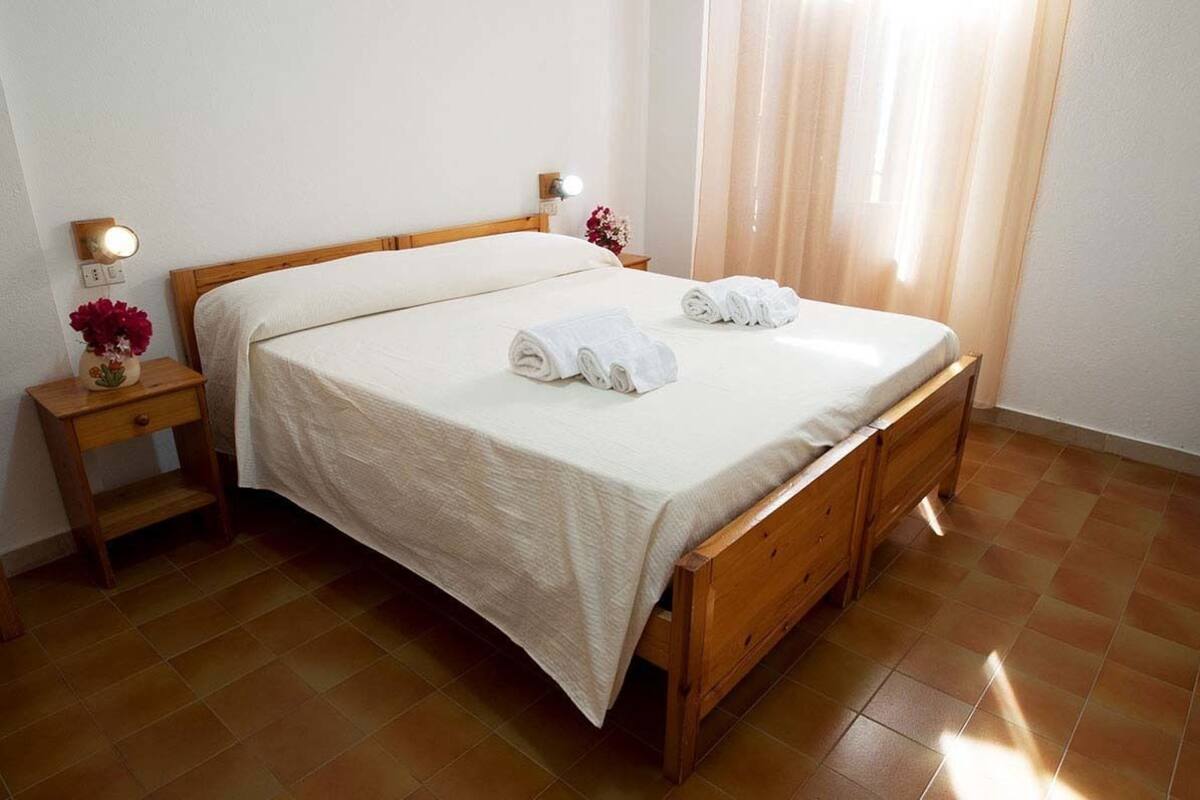 A double bed with a white bedspread and two rolled towels on the side. There is a wooden nightstand with a lamp and a vase of flowers. The room has a tiled floor and a window with sheer curtains.