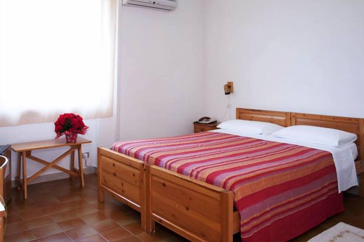 A bedroom with a wooden bed, a bedside table, a small nightstand, a red flower vase, and a window with blinds.