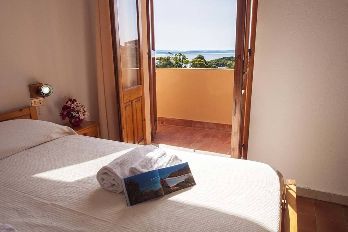A hotel room with a bed, a nightstand with a lamp, a window with a view of the sea, and a door leading to an outdoor balcony.