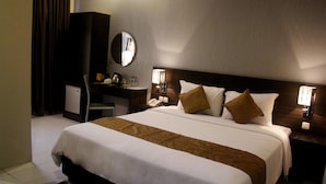 A hotel room with a large bed, a desk, a chair, a mirror, and a nightstand with a lamp.