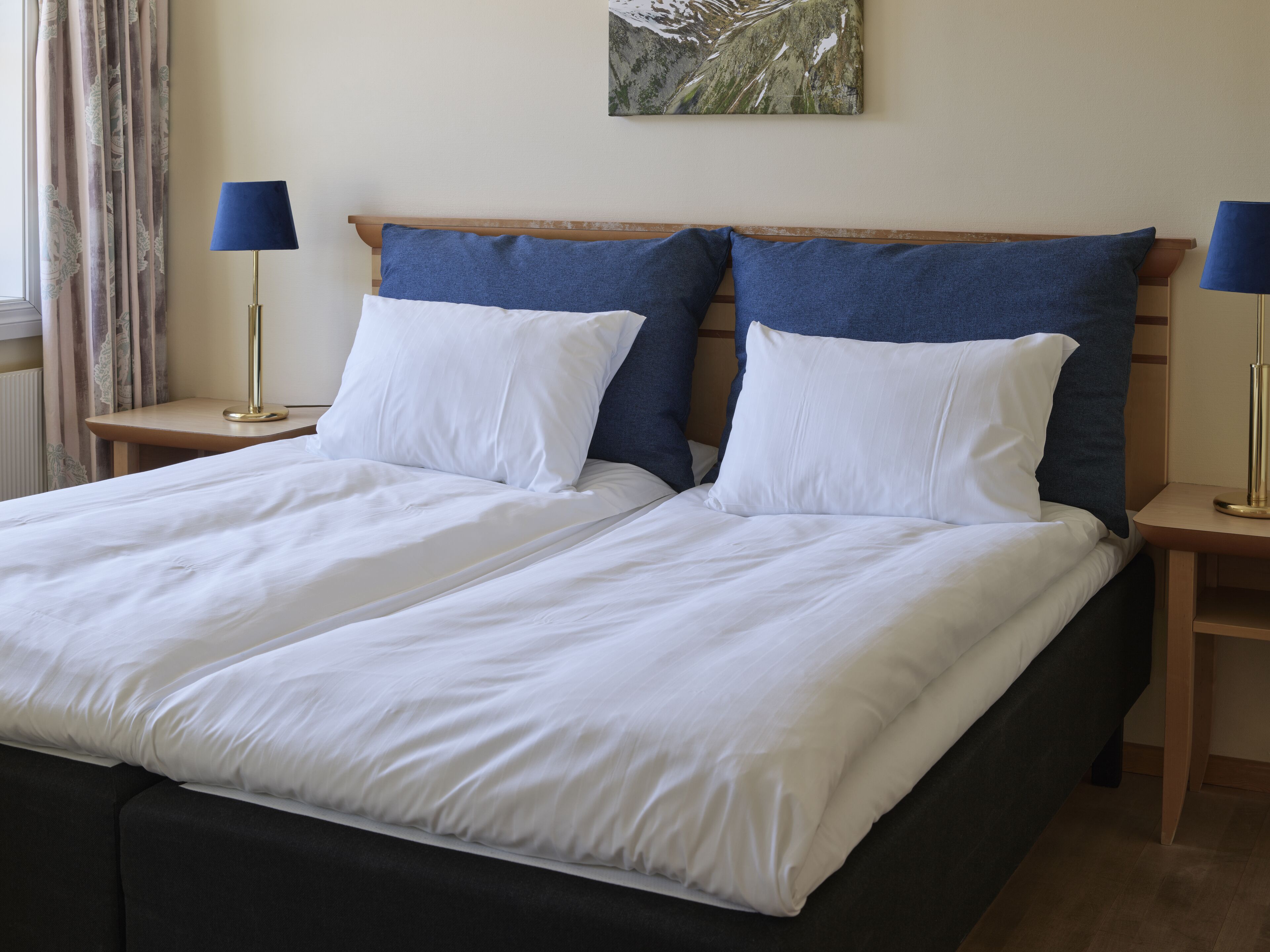 A double bed with white linens, two blue pillows, and a wooden headboard. There are two bedside tables with lamps and a painting on the wall.